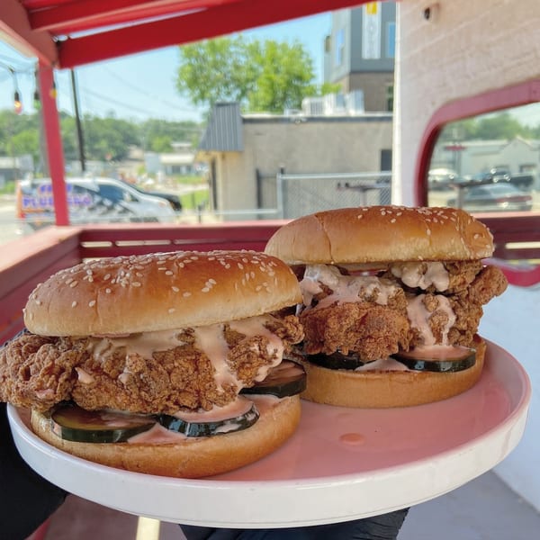 Two crispy chicken sandwiches with sauce and pickles on a pink plate, outdoors at Picone.