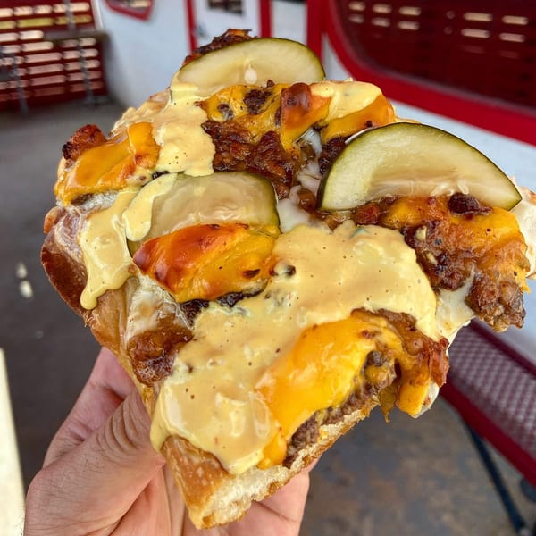 A hand holds a slice of Picone's Italian pizza topped with melted cheese, meat, and pickles.