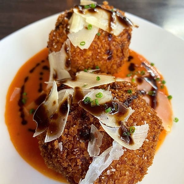 Two golden-brown arancini, drizzled with balsamic glaze and topped with parmesan shavings and chives.